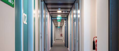 A long, narrow hallway with doors labeled 02 and 03, a fire extinguisher on the right, and emergency exit signs at the end.