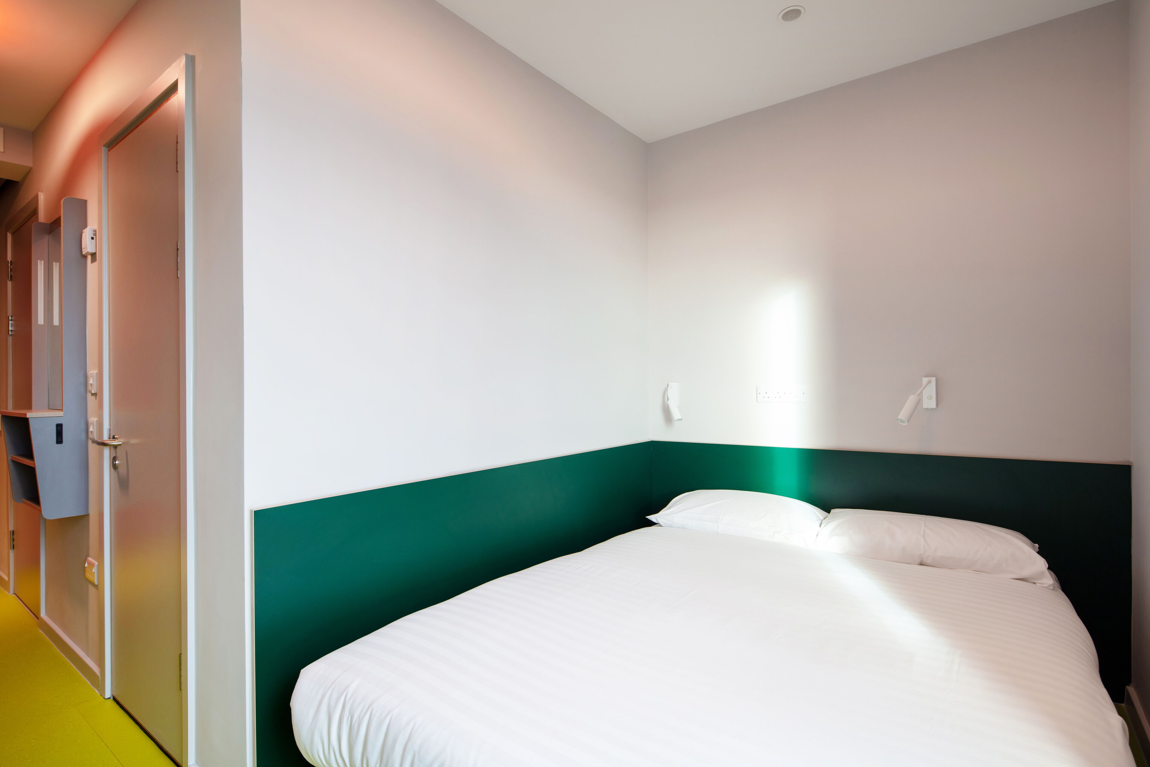 Habitación de hotel con una cama individual, cabecera verde y pared blanca.