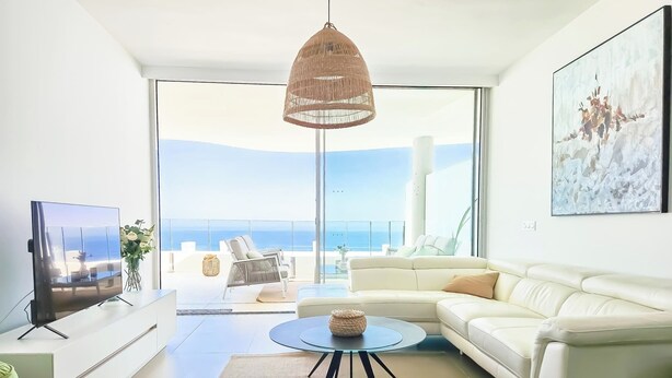 Ein modernes Wohnzimmer mit einem weißen Sofa, einem runden Glastisch und einem großen Fernseher. Ein Balkon bietet Blick auf das Meer.