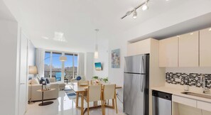A modern kitchen with a stainless steel refrigerator, a dining area with a table and chairs, and a large window offering a city view. - Pool + Infinity View + Balcony + Gym (Hallandale Beach)