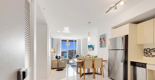 A modern kitchen with a stainless steel refrigerator, a dining area with a table and chairs, and a view of the city through large windows. - Pool + Infinity View + Balcony + Gym (Hallandale Beach)