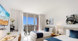Une chambre d’hôtel moderne avec deux lits, un balcon offrant une vue sur la ville, un bureau avec un ordinateur et des œuvres d’art accrochées aux murs.