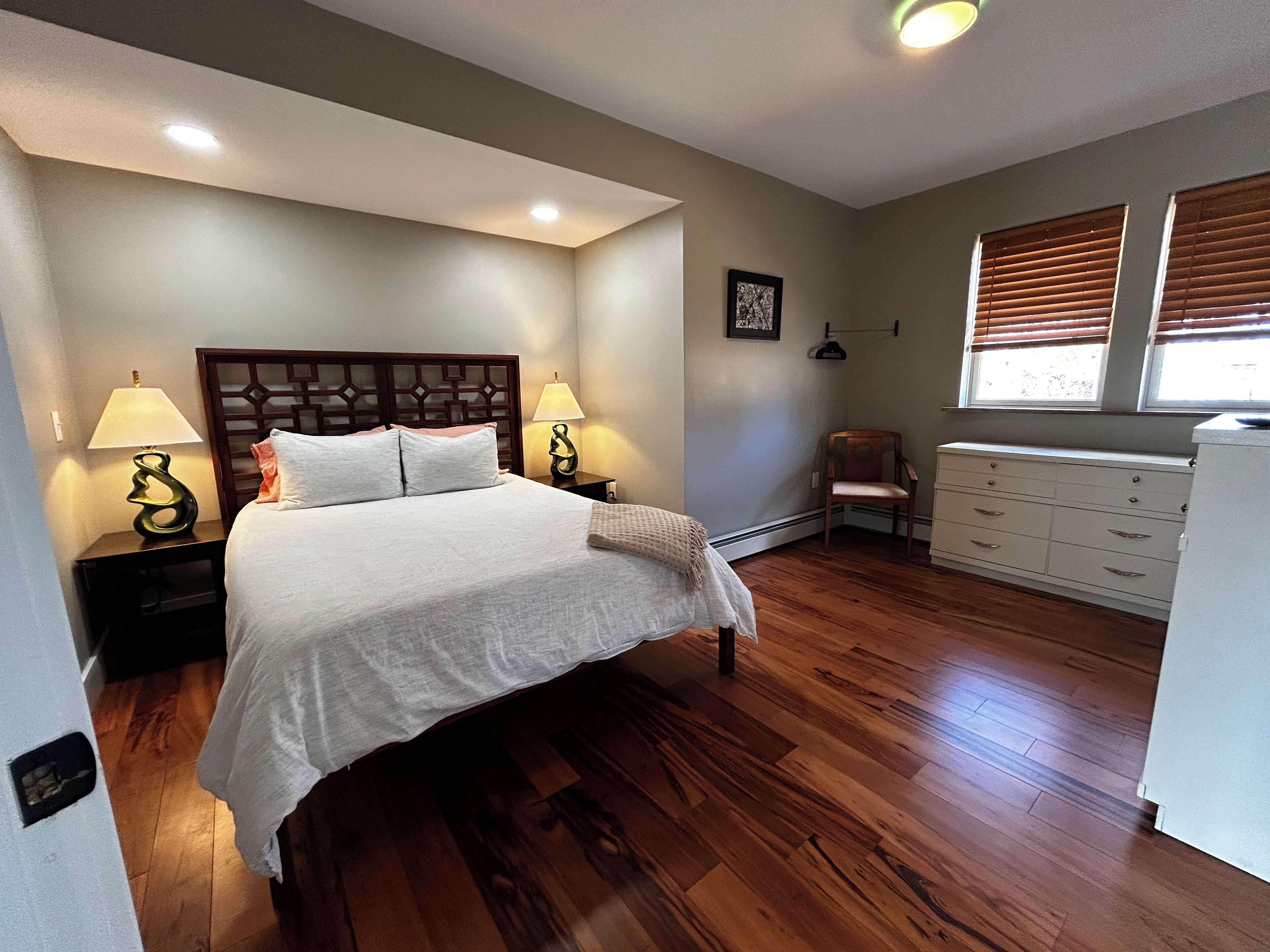A bedroom with a wooden bed, two bedside tables with lamps, a chair, a dresser, and a window with blinds.