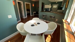 Een eetgedeelte met een ronde tafel en vier stoelen, een keuken op de achtergrond, en uitzicht buiten door een raam.