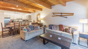 Ein gemütliches Wohnzimmer mit Holzbalkendecke, einer Sofagarnitur, einem Couchtisch und einer Küche im Hintergrund.