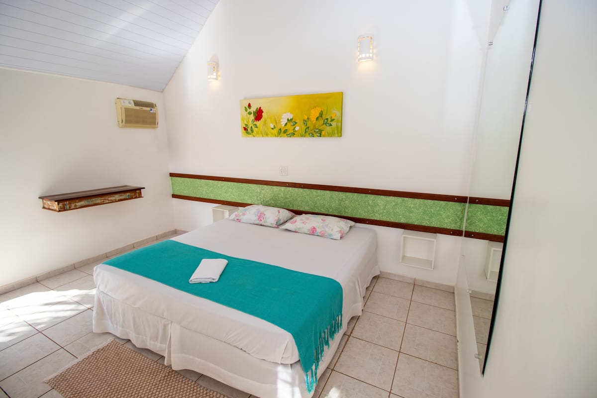 A bedroom with a bed, a painting, a shelf, and a wall-mounted air conditioner.