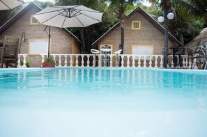 A swimming pool with a white balustrade, a large umbrella, and a brick building with windows and doors in the background.