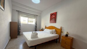 Een slaapkamer met een bed, een nachtkastje, een dressoir, een raam met gordijnen en een schilderij aan de muur.