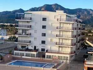 Un hotel de varios pisos con balcones, piscina y montañas al fondo.
