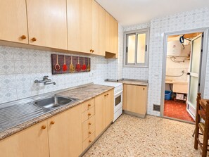 A compact kitchen with wooden cabinets, a stainless steel sink, a stove, and a window with a view of the outside. - WIMPY esc.B 1º  Solo Familias! (Xeraco Playa)