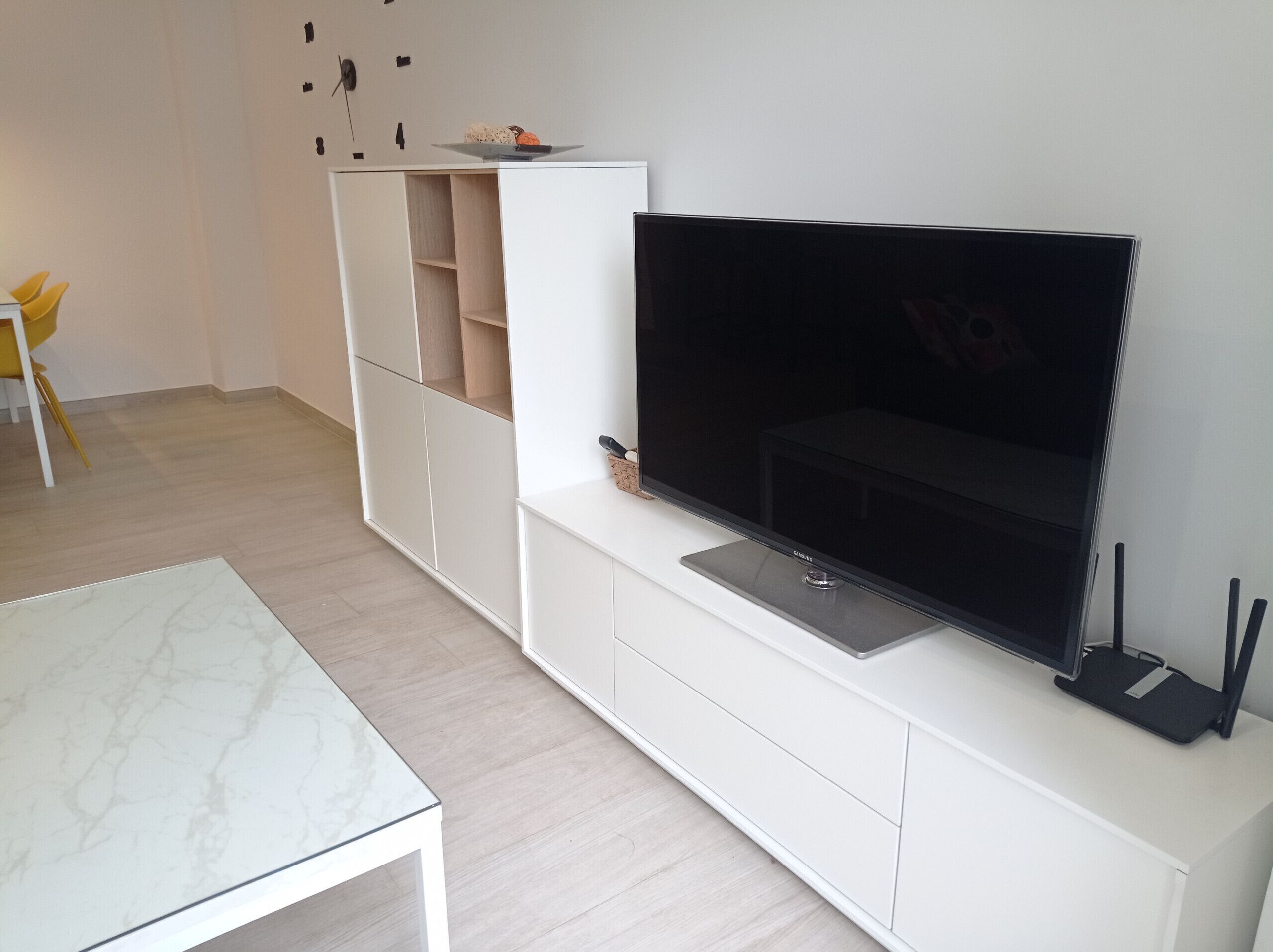 A modern living room with a flat-screen TV mounted on a white entertainment unit, a small table, and a yellow chair.