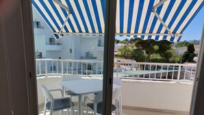 A balcony with white plastic chairs and a table, overlooking a pool area with a blue and white striped awning.