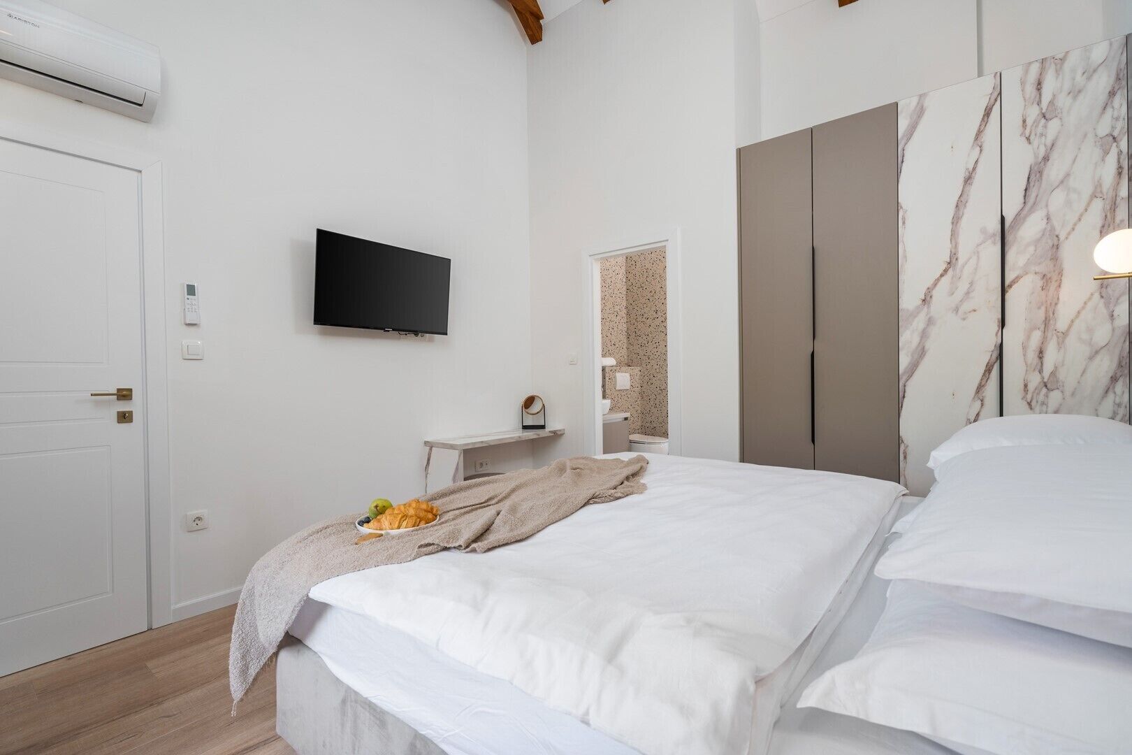 A bedroom with a bed, a TV mounted on the wall, a wardrobe, and a bathroom visible through an open door.