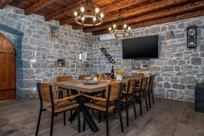 A stone-walled dining area with a wooden table and chairs, a chandelier, and a mounted television.