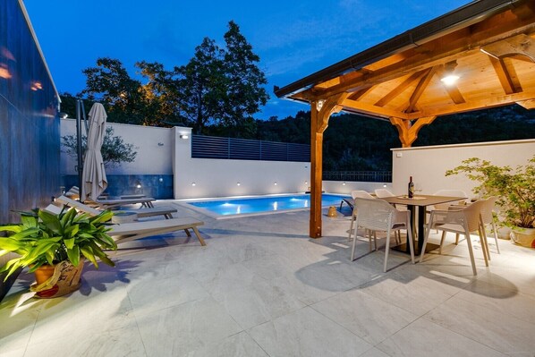 A modern outdoor pool area with a pergola, lounge chairs, and a dining table.