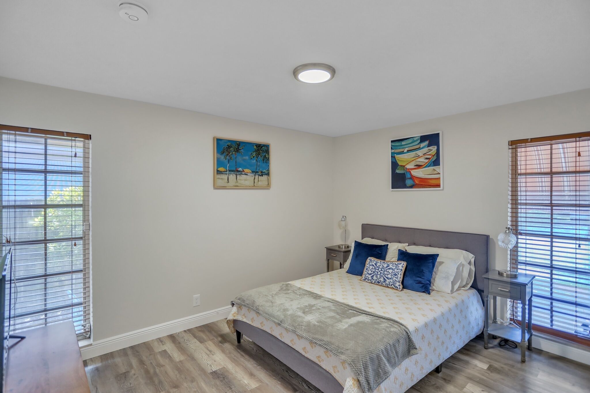A bedroom with a bed, two paintings, a nightstand, and a window with blinds.