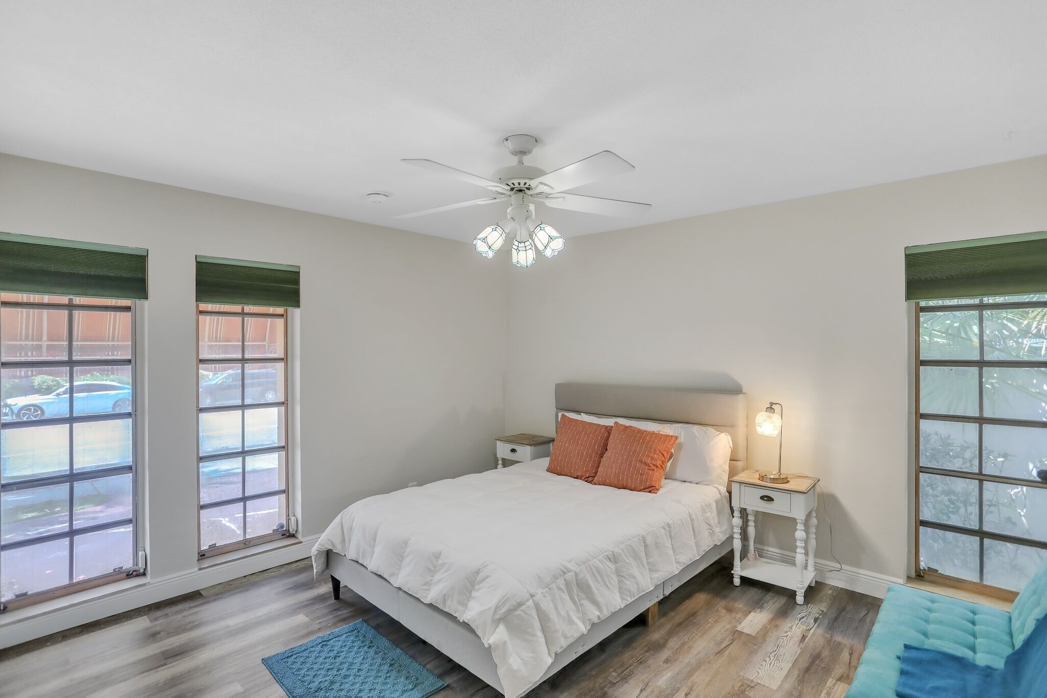 Une chambre à coucher comprenant un lit, des tables de chevet, un ventilateur de plafond et de grandes fenêtres équipées de stores.