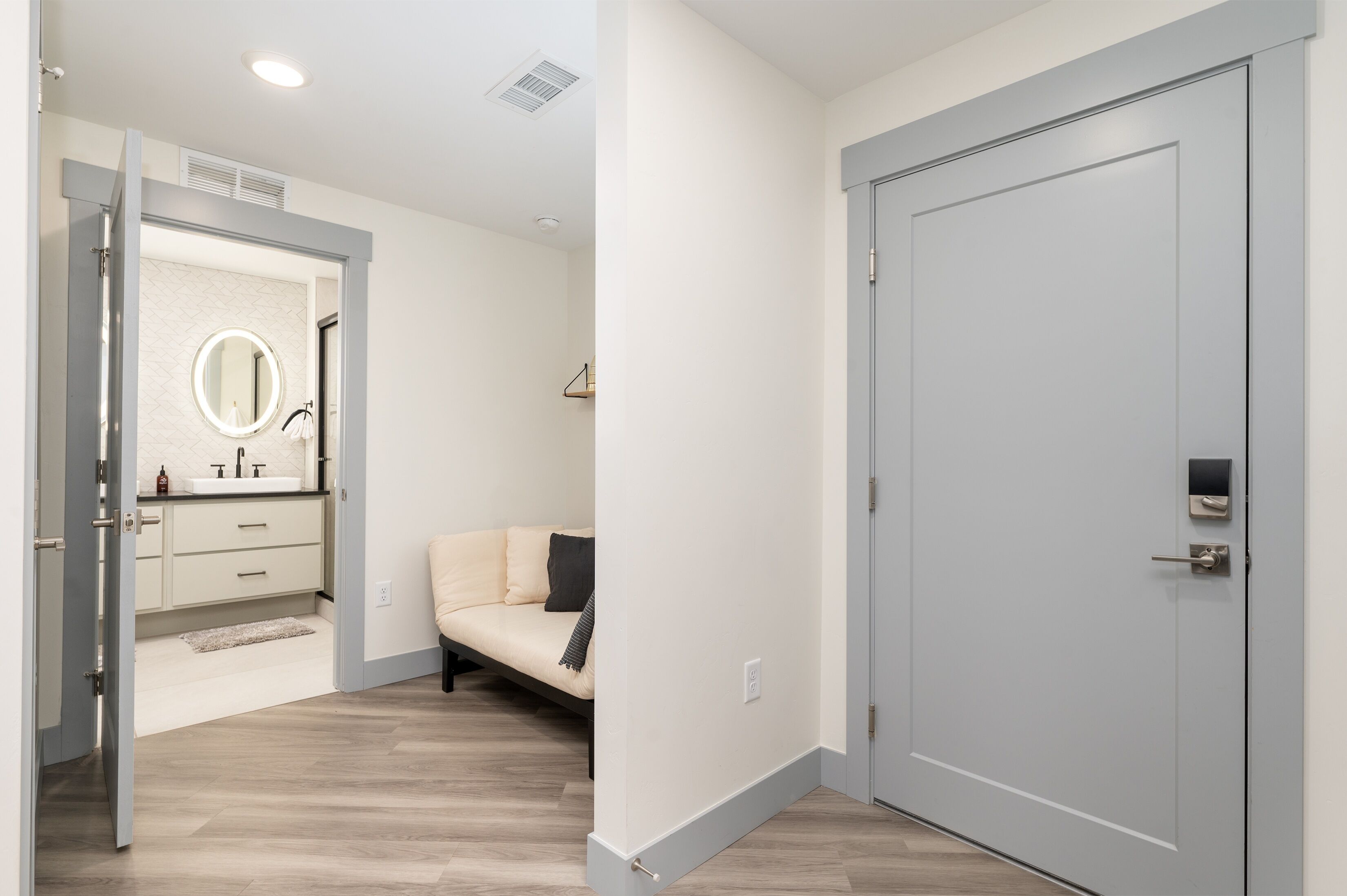 A modern bathroom with a round mirror, a sink, and a toilet. There is a beige sofa in the adjacent room.