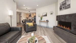 A modern living room with a fireplace, a sofa, a coffee table with a plant, and a kitchen in the background.