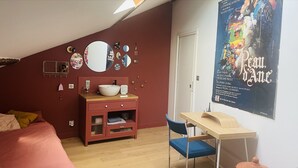 Een slaapkamer met een schuine plafond, een bed, een bureau met stoel, een dressoir, een spiegel en een poster aan de muur.