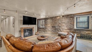 A spacious living room with a stone wall, leather sofa, fireplace, and a flat-screen TV. - Modern 2BR Steps from Main St! (Park City)