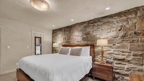 A bedroom with a stone wall, a wooden bed, a bedside table with a lamp, and a bench. - Modern 2BR Steps from Main St! (Park City)