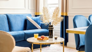 Un canapé bleu avec un coussin blanc, une table ronde sur laquelle se trouve une assiette de croissants, un verre de jus d’orange, un vase noir orné de feuilles dorées et une petite tasse de café.
