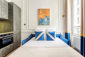 A hotel room with a bed, a painting, a modern kitchen, and a view of a building.