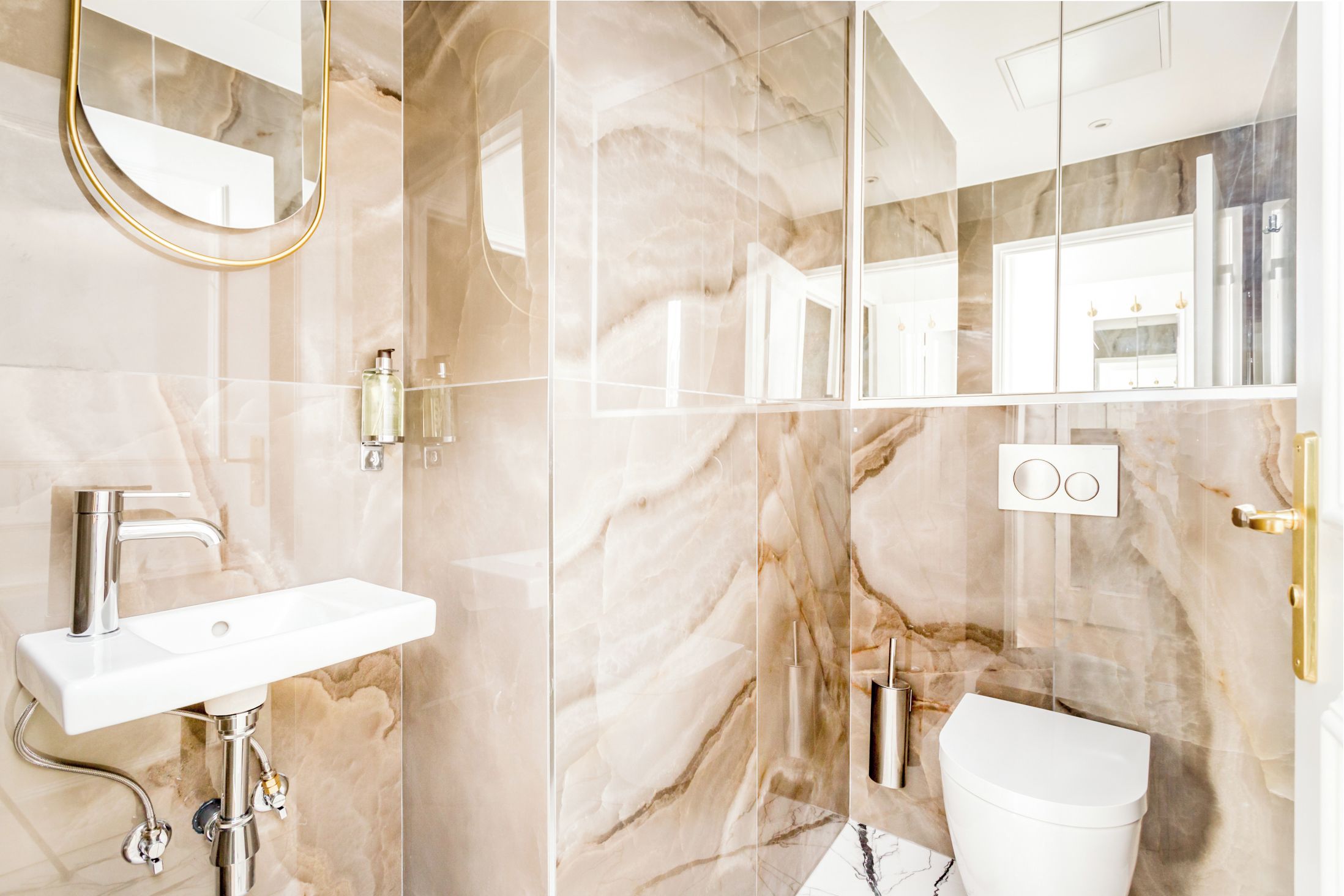 A modern bathroom with a marble backsplash, a white sink, a toilet, and a gold-framed mirror.