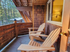 Eine Holzterrasse mit Adirondack-Stühlen, ein Fenster mit Lampe und Blick ins Grüne.