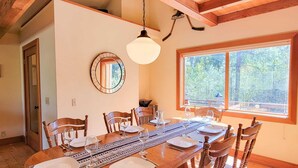 Une salle à manger avec une table en bois dressée pour six personnes, un luminaire suspendu, un miroir et une fenêtre donnant sur des arbres.