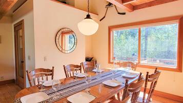 Un comedor con una mesa de madera dispuesta para seis personas, una lámpara colgante, un espejo y una ventana con vistas a árboles.