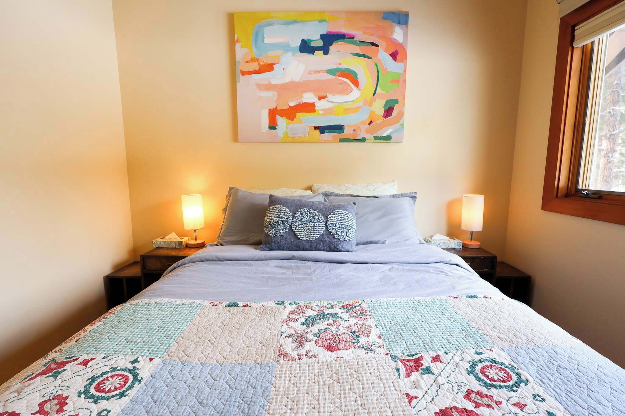 A bed with a quilt, two bedside lamps, a wall art piece, and a window.