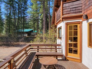 Eine Holzterrasse mit Bänken und einem Tisch, ein Gebäude mit einem Holzbalkon und bewaldete Umgebung.
