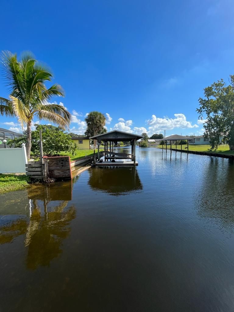 A calm water body with a dock and a covered structure, surrounded by greenery and clear skies.