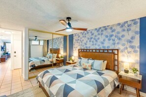 A bedroom with a large bed, a ceiling fan, a nightstand with a lamp, and a mirror.