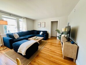 Un salon avec un canapé d’angle bleu foncé, une table basse en bois, un téléviseur posé sur un meuble en bois et une grande fenêtre avec des rideaux.