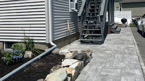 Un edificio residencial con fachada gris, una escalera visible que lleva a la entrada y un camino pavimentado con borde de piedra.