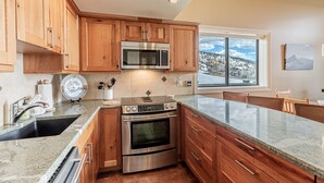 A kitchen with wooden cabinets, a stainless steel microwave, an oven, and a large window offering a view of a snowy landscape. - Ski-In/Ski-Out Condo with Sweeping Slopeside Views! (Steamboat Springs)