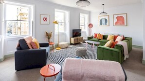 A modern living room with a sofa, armchair, and a coffee table. There are two windows with city views, a floor lamp, and a TV on a wooden stand. - Stunning apartment in central location - Pass the Keys (Bath)