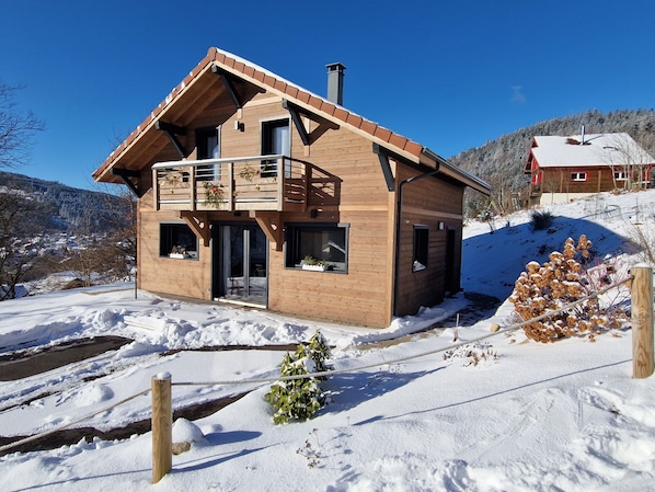 A wooden chalet with a balcony, a chimney, and a view of a snowy landscape.