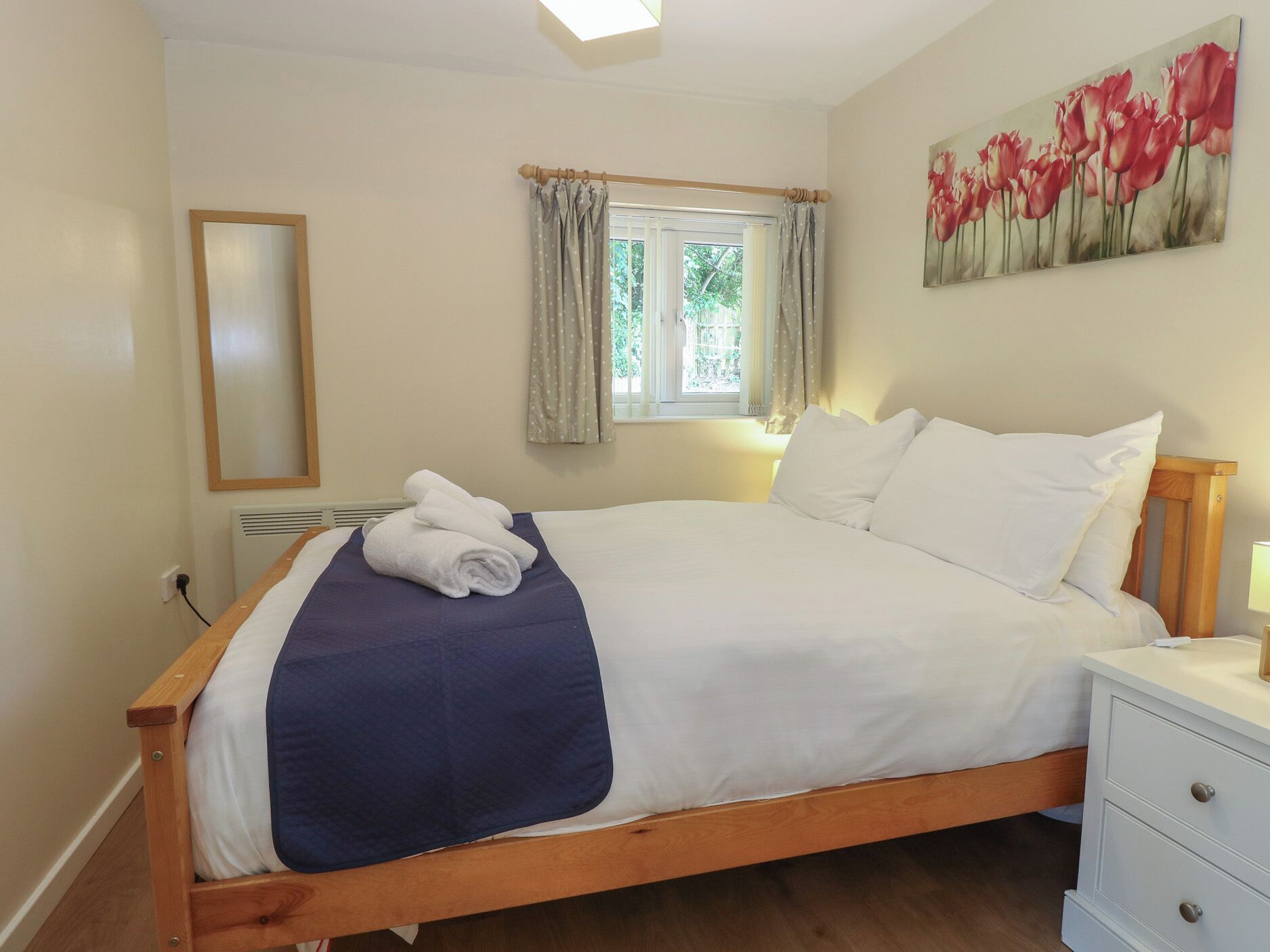 A bedroom with a bed, a bedside table, a nightstand, a window with curtains, a wall-mounted mirror, and a painting of tulips.