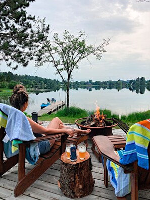 Een terras aan het meer met houten meubels, een vuurkuil en uitzicht op een rustig meer en gebouwen in de verte.