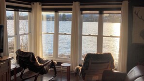 Un salón acogedor con un ventanal grande que da a un lago, amueblado con sofás de cuero y una mecedora.