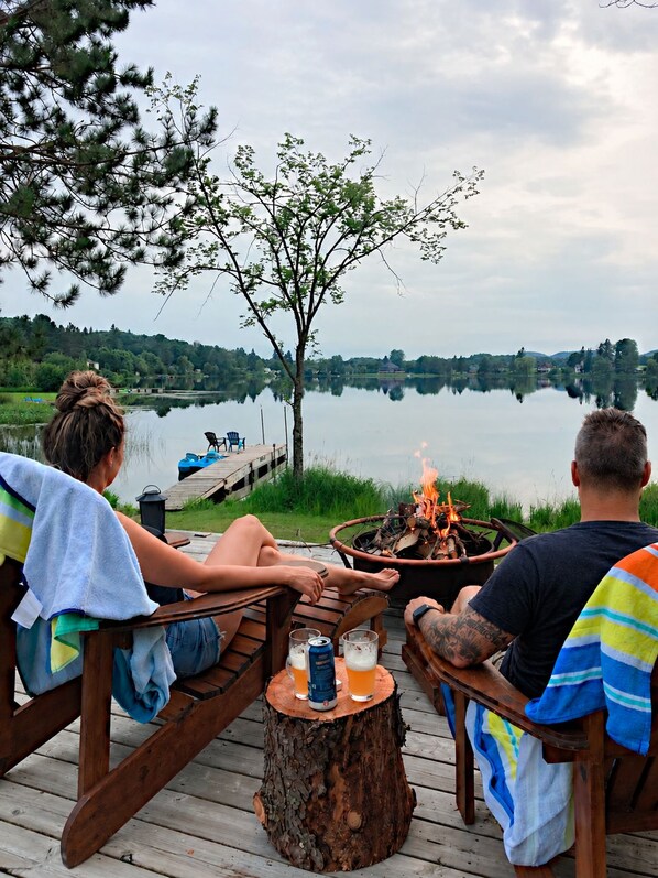 Een houten terras met een vuurkuil, twee personen zittend, en uitzicht op een meer.