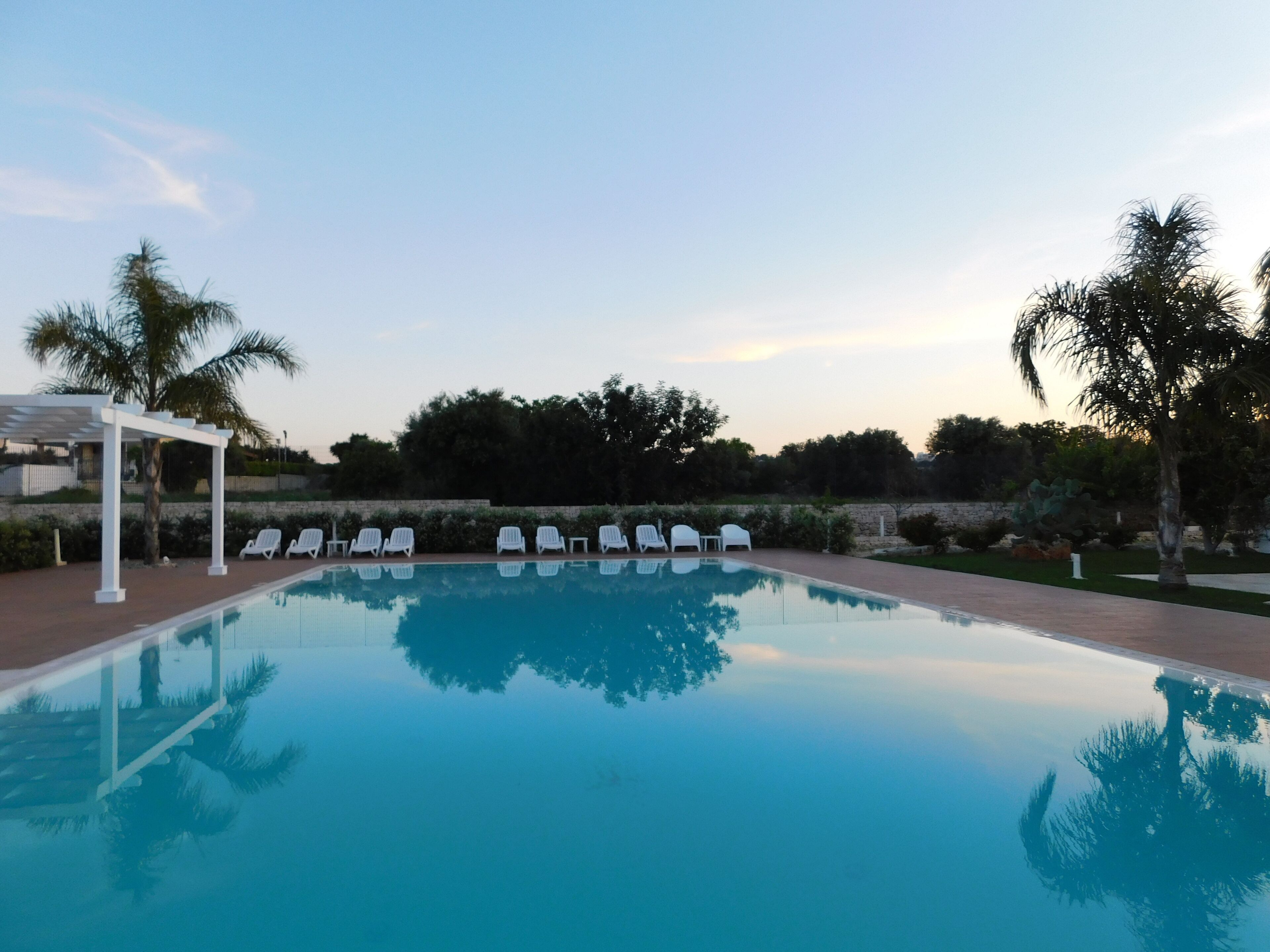 A swimming pool with white lounge chairs and a pergola, surrounded by palm trees and greenery.