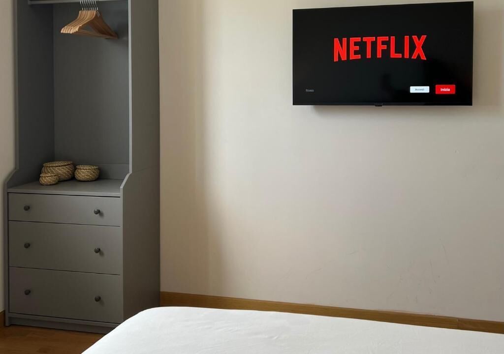 Une chambre avec une commode grise, un porte-manteau en bois suspendu, un téléviseur à écran plat fixé au mur qui affiche le logo « NETFLIX », et un lit avec une literie blanche.