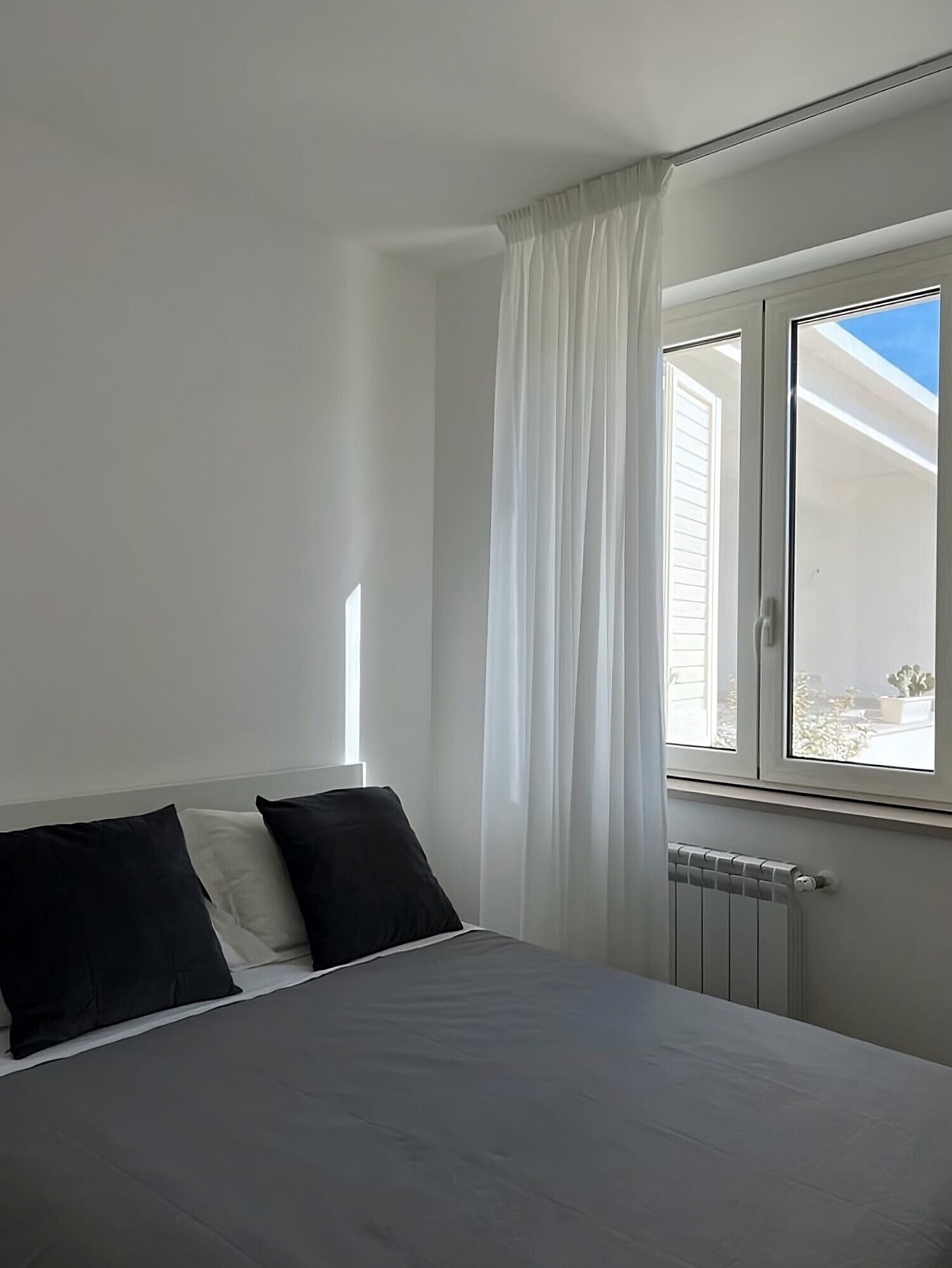 Ein Schlafzimmer mit einem Bett, weißen Vorhängen, einem Fenster und einer Heizung.