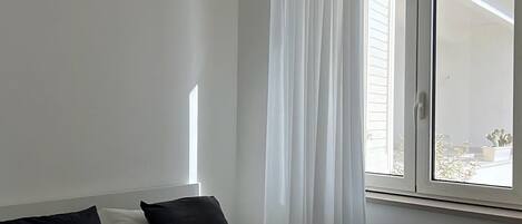 Ein Schlafzimmer mit einem Bett, weißen Vorhängen, einem Fenster und einer Heizung.