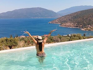 A person in a swimming pool with arms raised, overlooking a coastal landscape with mountains and a bay. - Augustela Villa Seaview Vacation Villa (Kefalonia)